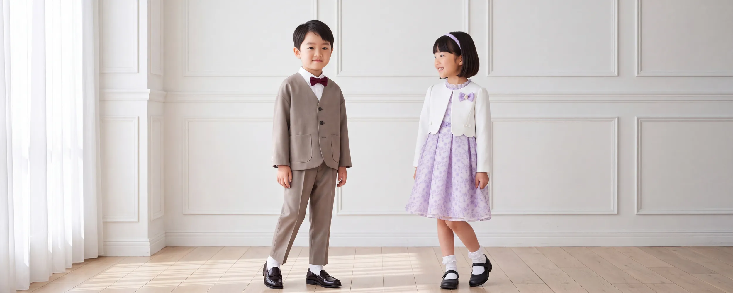 ベージュのジャケットとパンツに蝶ネクタイを合わせた男の子と、白いボレロに薄紫のワンピースを合わせた女の子が並んで立つ様子